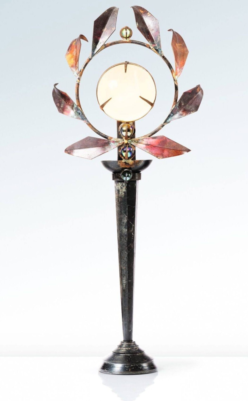 Decorative metal sculpture with leaf-like details on a tall stand, featuring a circular center piece against a light background