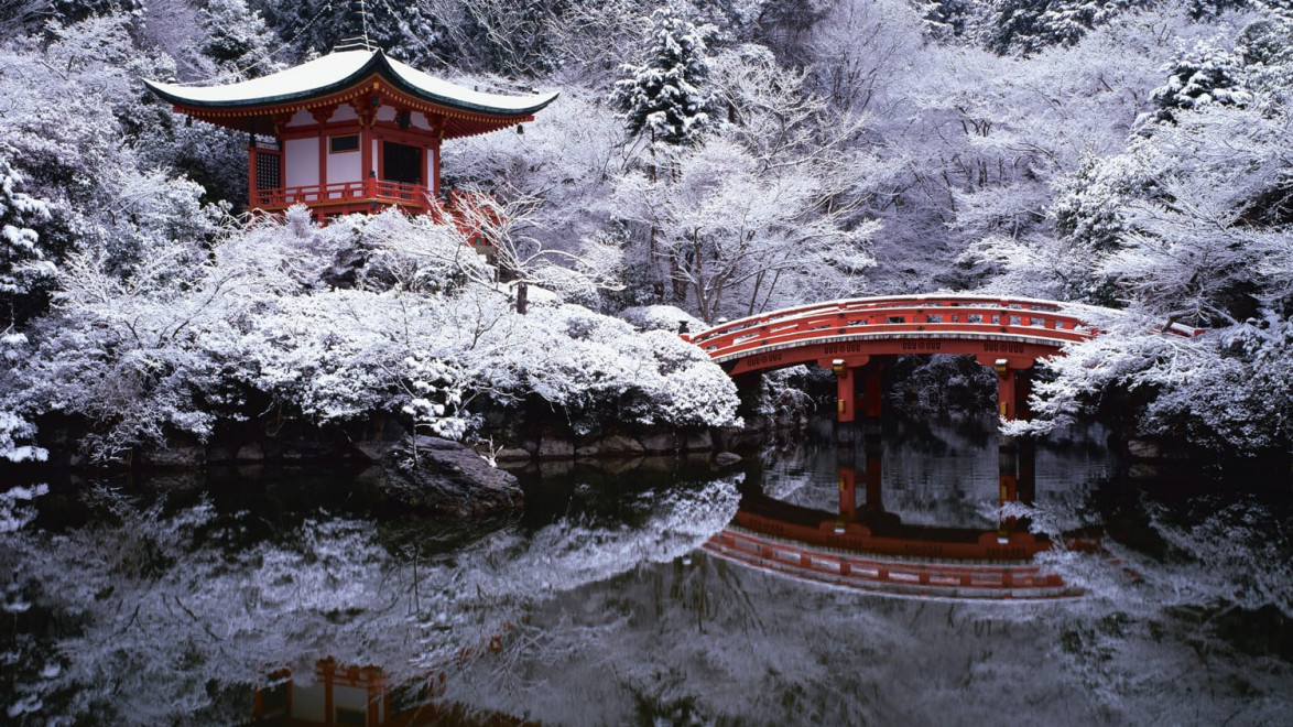 Snow-covered Japanese temple with red bridge reflected in still water, surrounded by snowy trees and mountains.