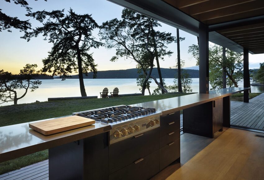 Outdoor kitchen with stovetop and cutting board overlooking a serene lake view at sunset, trees and deck visible.