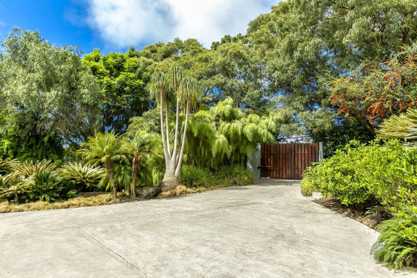 Driveway leading to a wooden gate surrounded by lush greenery and tall trees under a partly cloudy sky.