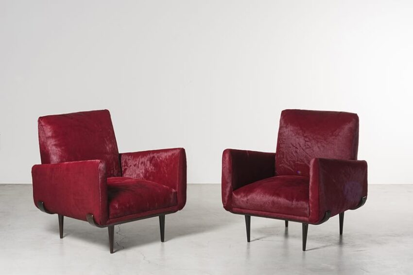 Two vintage red velvet armchairs with wooden legs on a light gray floor against a white wall.