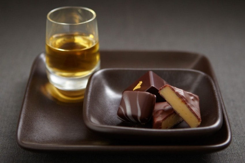 Glass of whiskey with assorted chocolates on a dark ceramic plate.