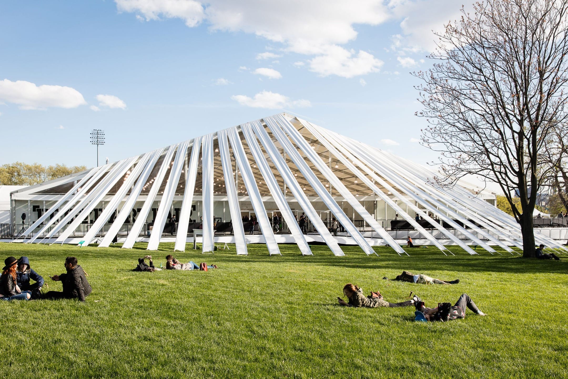 Modern outdoor pavilion with white beams, large open space surrounded by grass, people lounging on the lawn, and a clear sky above.