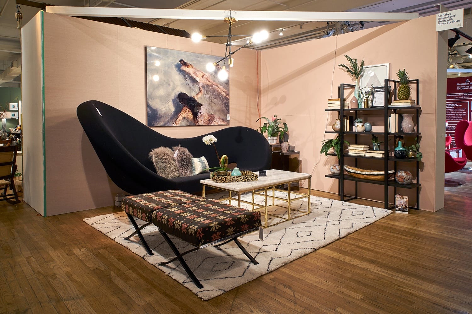 Modern living room with a black sofa, patterned bench, artwork, and shelving on a pink wall, decorated with plants and decor items.