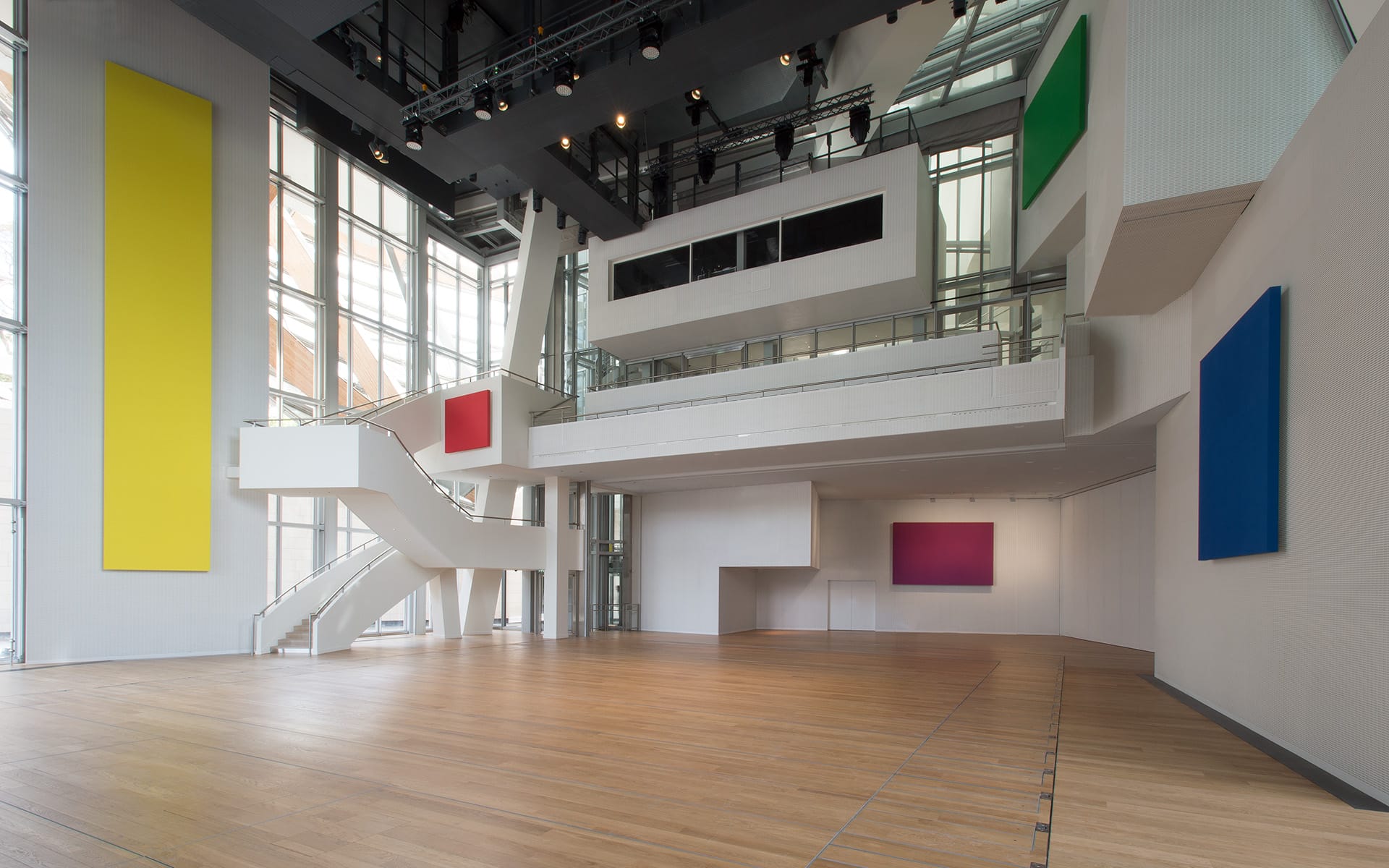 Modern gallery interior with large colorful abstract paintings on walls and wooden floors, featuring staircase and high ceilings.