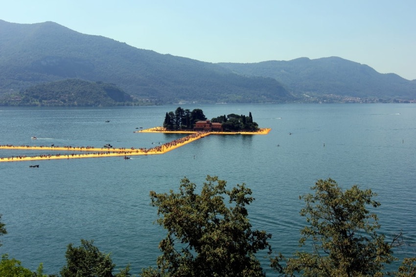 Floating walkway on a lake leading to a small island with trees and mountains in the background.
