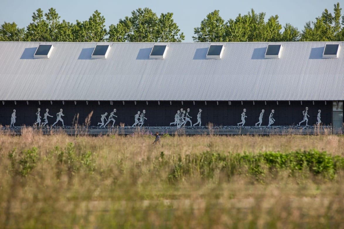 A mural of silhouetted figures walking on the side of a modern building, with tall grass in the foreground.