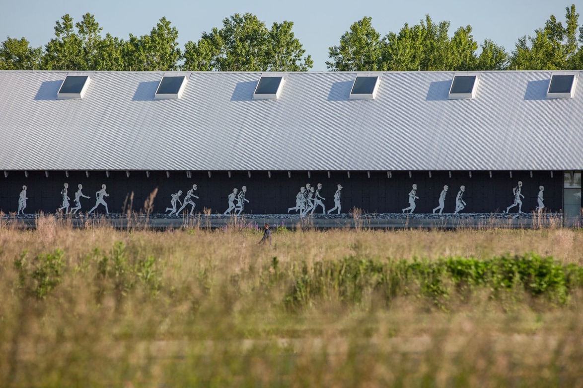 A mural of silhouetted figures walking on the side of a modern building, with tall grass in the foreground.