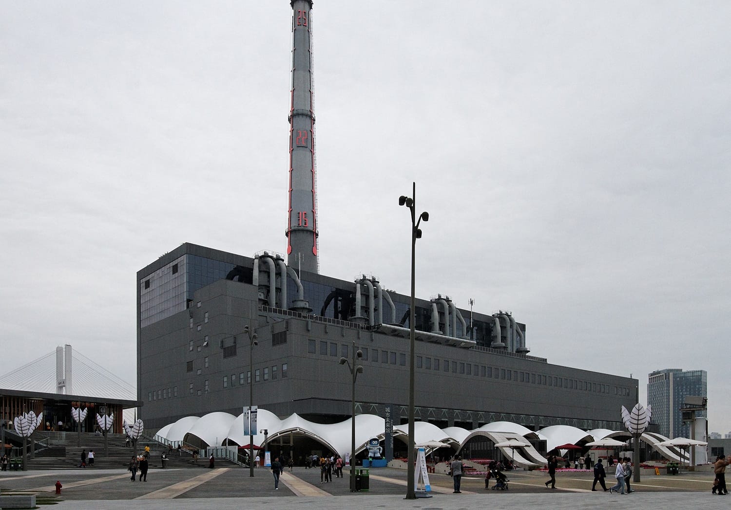 Large industrial building with smokestack and modern entrance, people walking nearby, cloudy sky in the background.