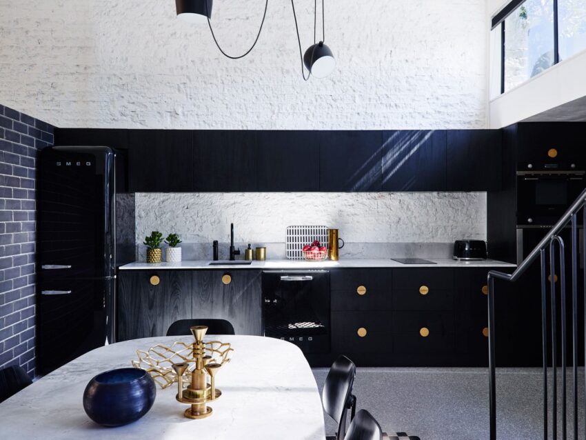 Modern kitchen with black cabinets, gold accents, marble table, and white brick walls, featuring pendant lighting and large windows.