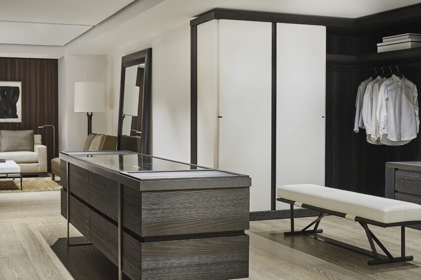 Modern walk-in closet with wooden flooring, white cabinets, open shelving with shirts, a bench, and a mirror.