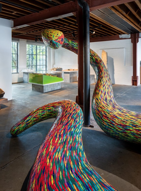 Colorful mosaic serpentine sculptures in an industrial art gallery setting with exposed beams and natural light from windows.