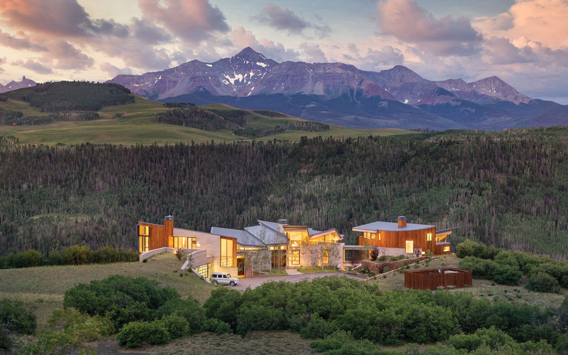 Modern mountain home with large windows and a scenic view of forested hills and distant snow-capped peaks under a cloudy sky.