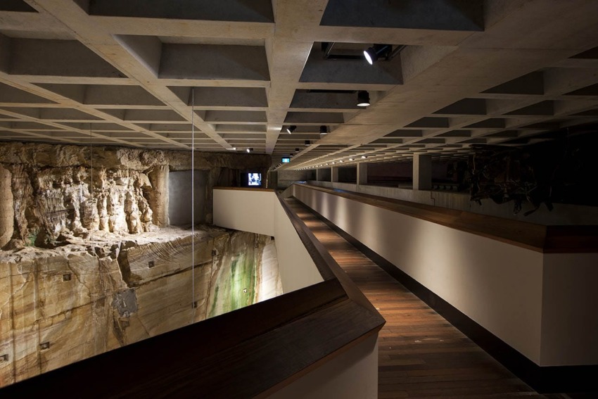 Interior view of a modern museum with wood and concrete elements, featuring a textured wall and wooden walkway.
