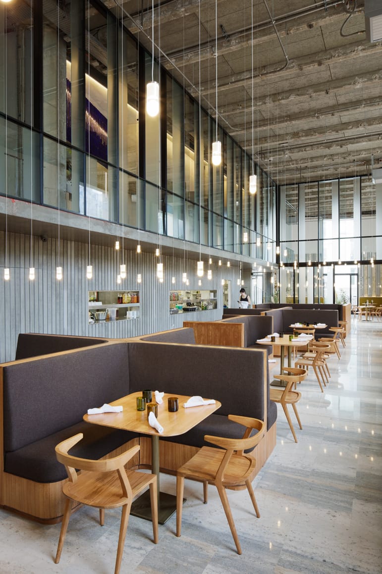Modern restaurant interior with wooden furniture, hanging lights, and large glass windows. Tables set with utensils and napkins.