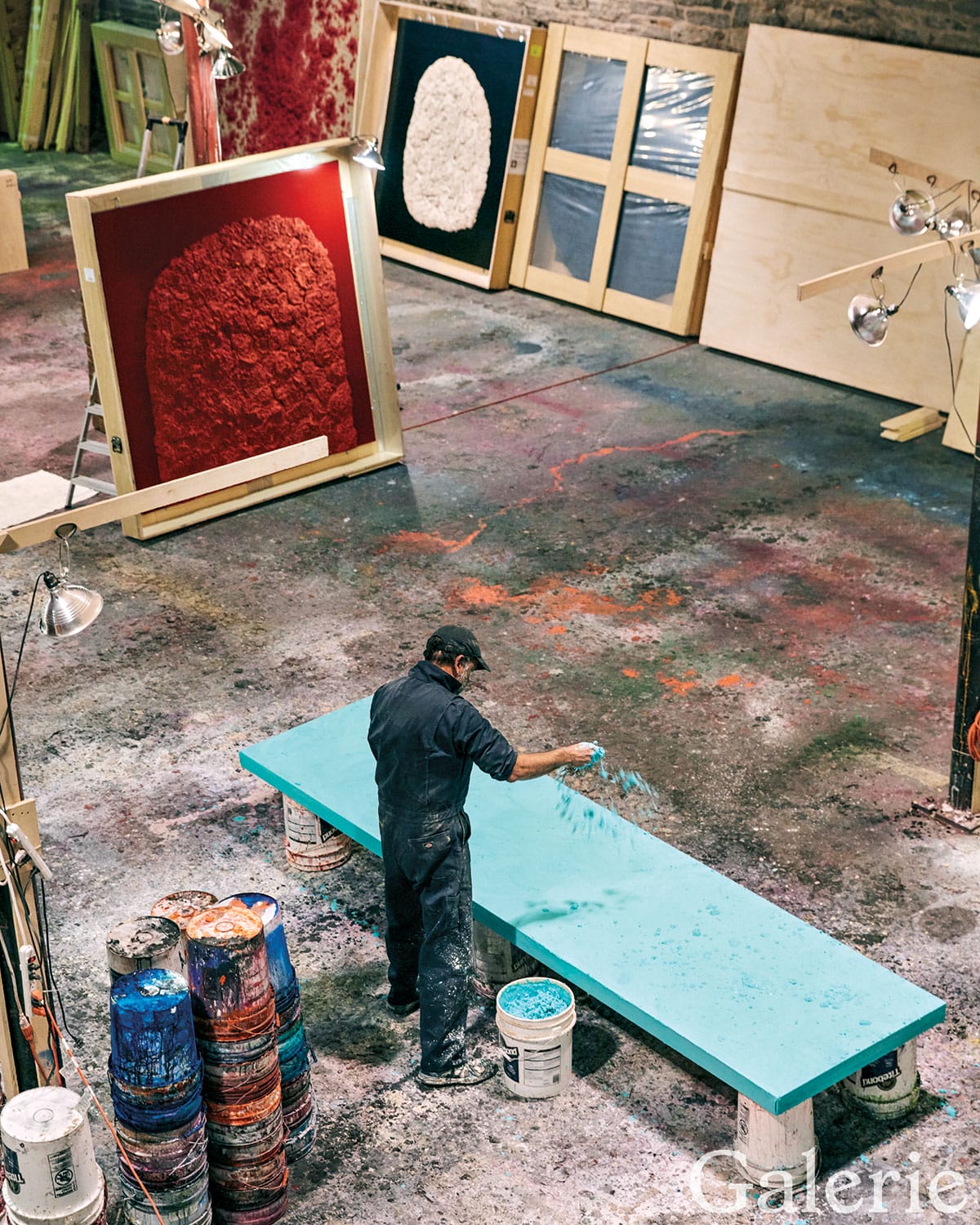 Artist working on a turquoise piece in a colorful studio with large textured artworks and paint buckets around.