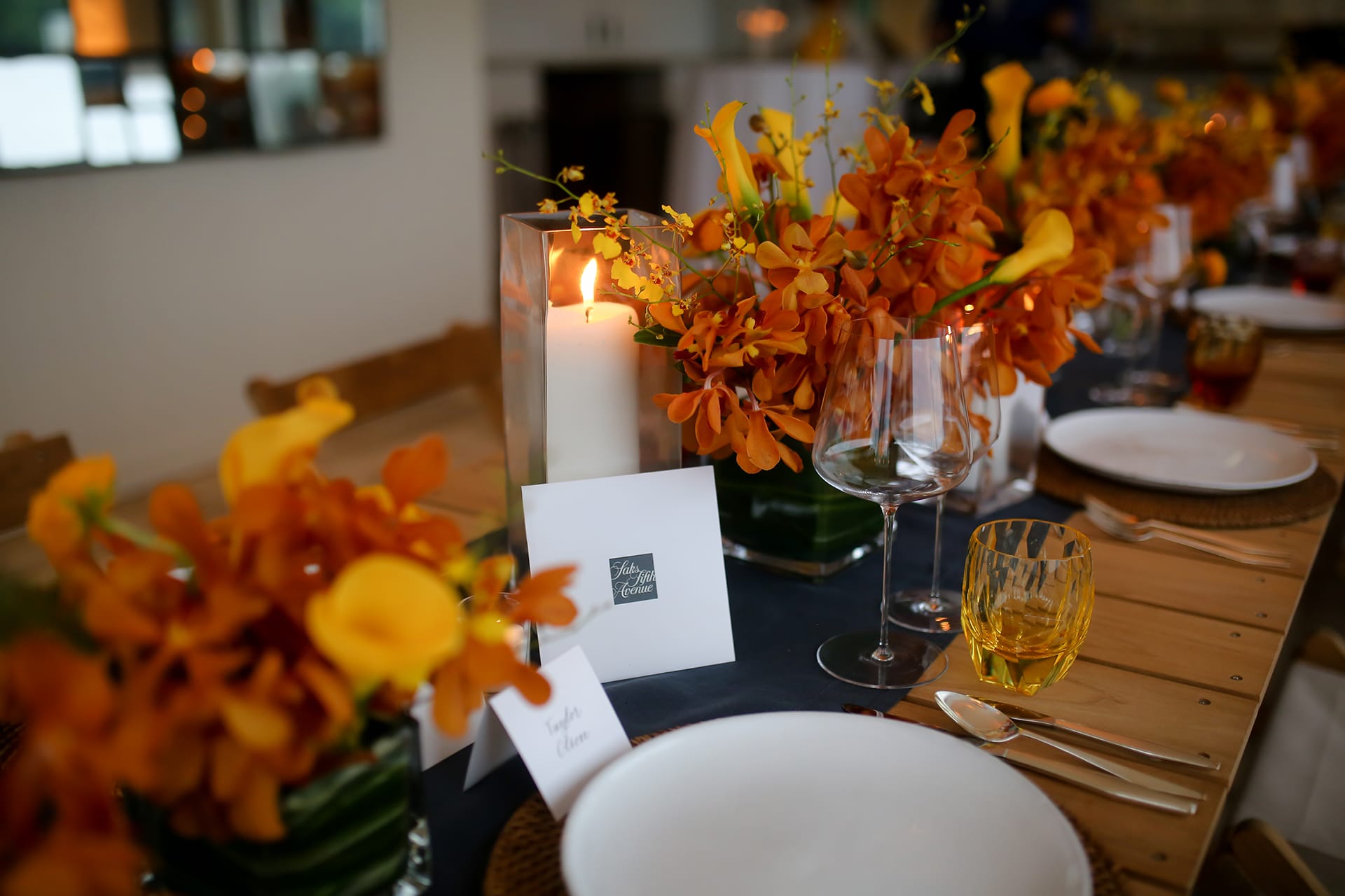 Table set with orange flowers, lit candle, wine glasses, and place cards at an elegantly decorated dinner event.