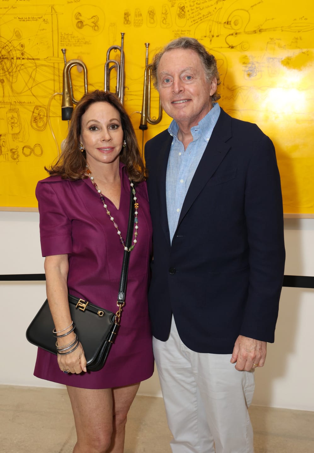 A woman in a purple dress and a man in a blazer standing in front of a yellow wall with instruments and drawings.