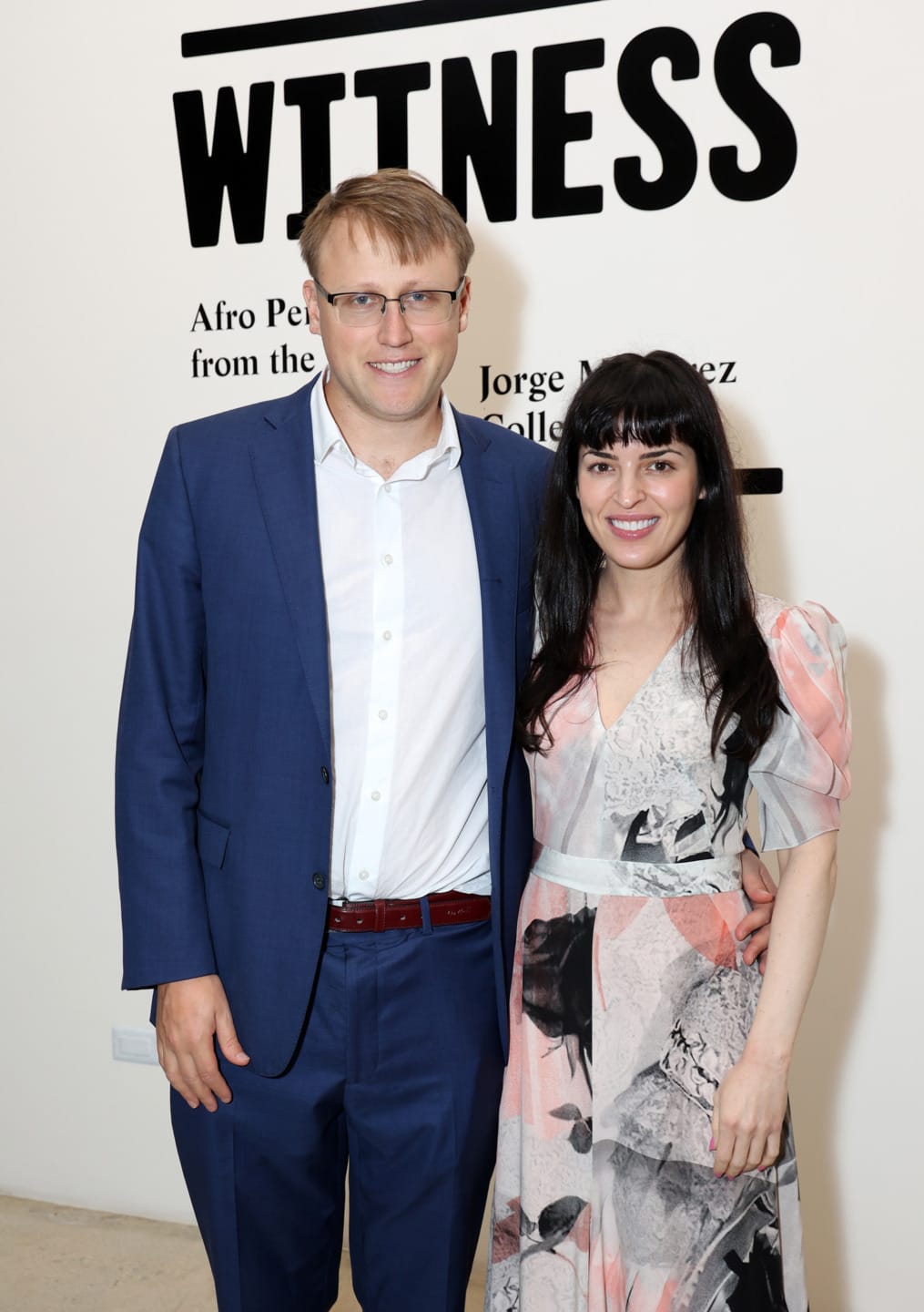 A man in a blue suit and a woman in a floral dress pose for a photo at an art exhibition.