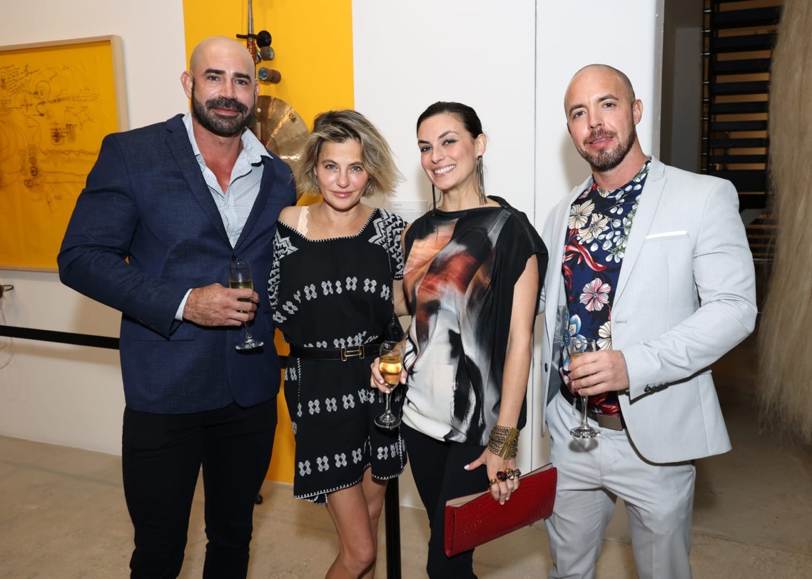 Group of four people dressed stylishly at an art gallery event, holding drinks and posing near colorful artwork on the wall.