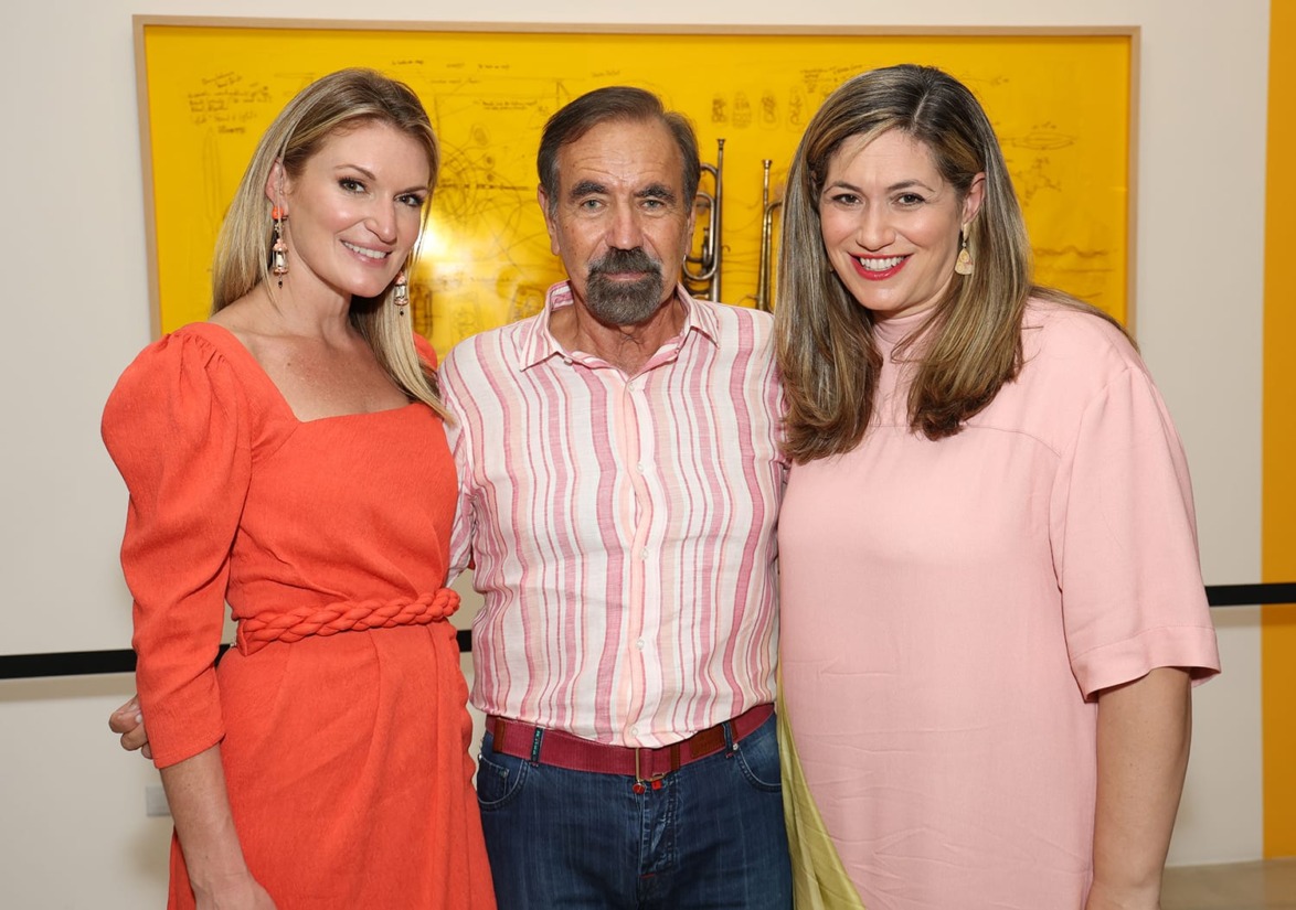 Three people standing together, smiling, with a yellow artwork in the background.