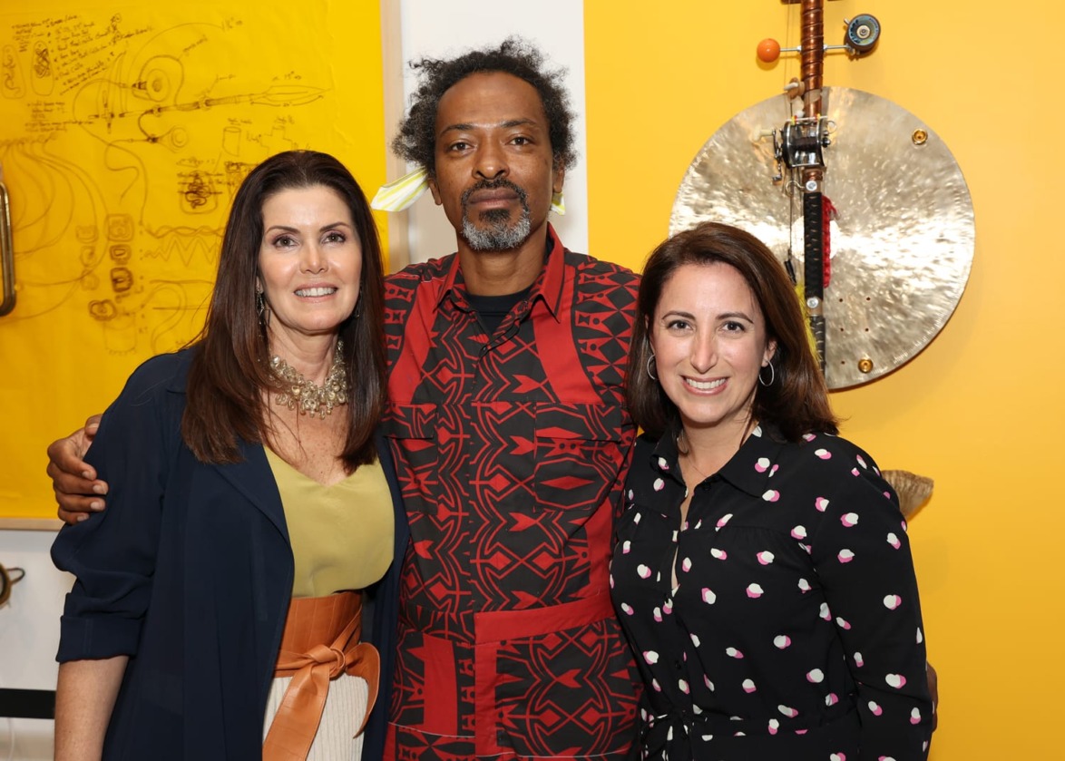 Three people posing together in front of vibrant artwork with yellow background.