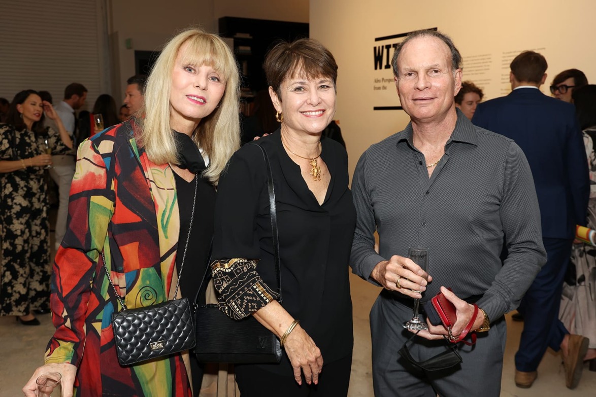 Three people smiling at a gallery event, with one holding a drink and another a red item.