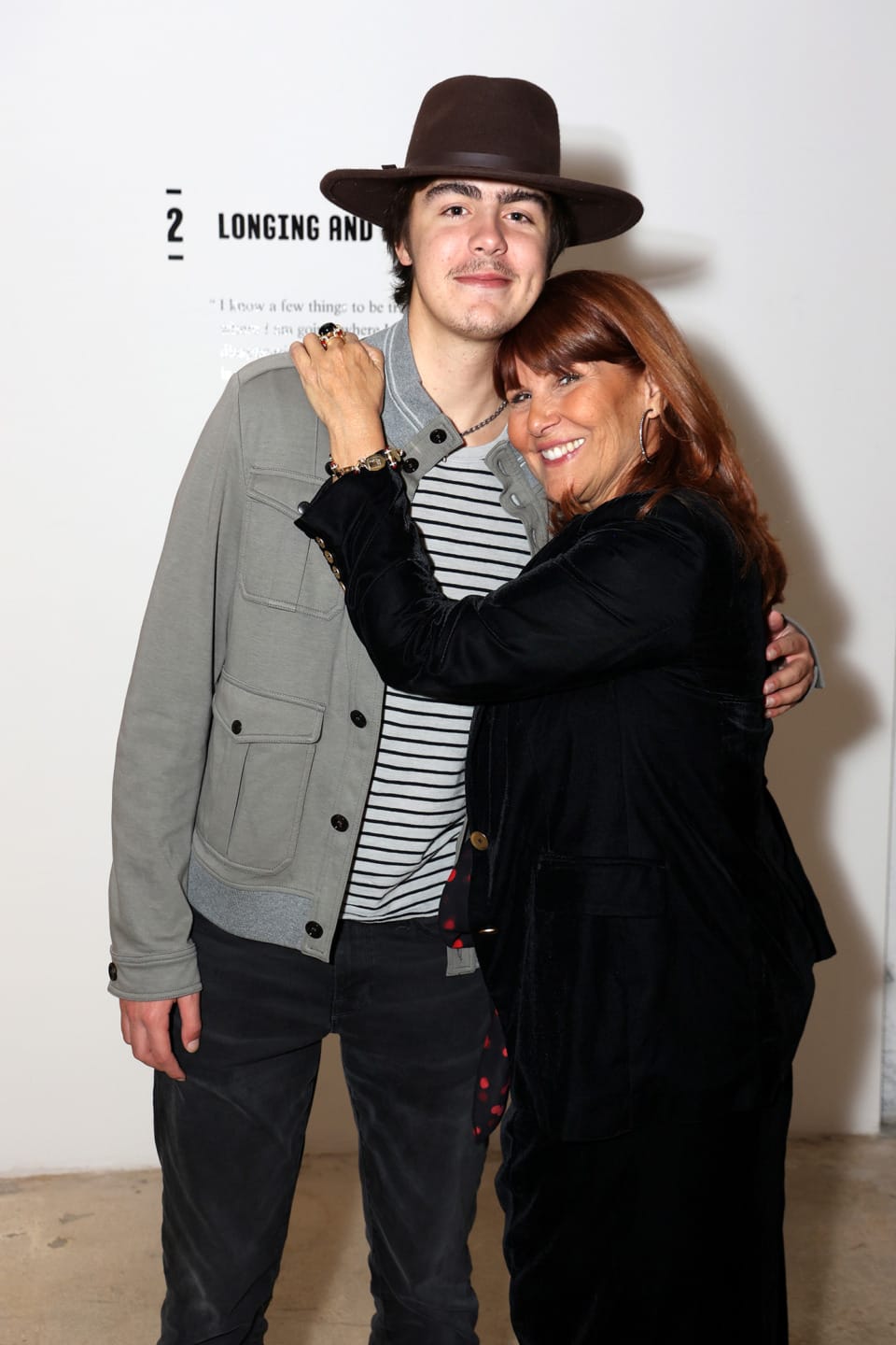 Woman smiling and embracing a young man wearing a hat in front of a wall with text.