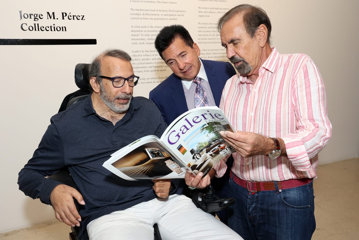 Three men stand together in an art gallery, one in a wheelchair, as they look at a magazine named "Galerie".