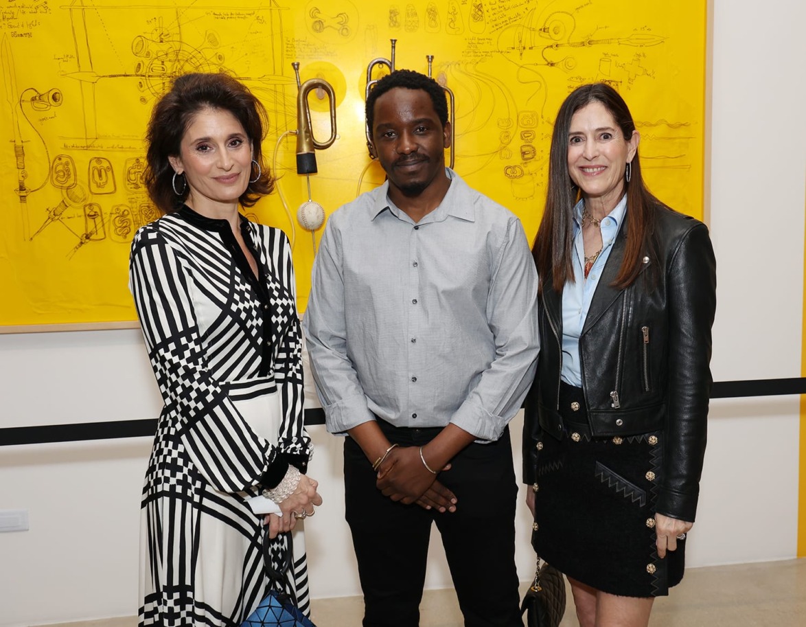 Three people posing in front of a yellow art piece with black sketches at an exhibition.