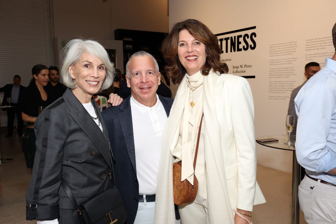 Three people smiling at an art gallery event, standing together with exhibition text on the wall in the background.