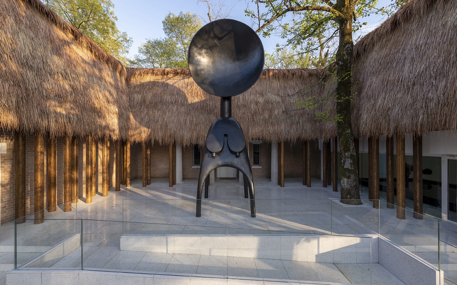Large black sculpture in courtyard with thatched roof buildings and trees in the background.