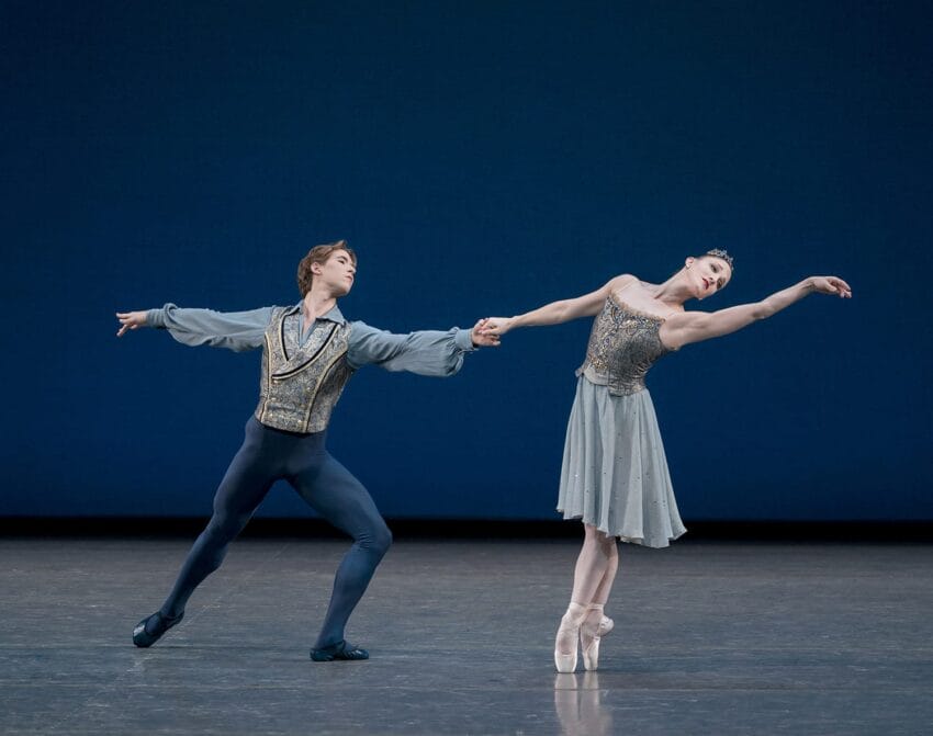 Ballet dancers performing on stage, holding hands gracefully, man extending arm, woman on pointe, deep blue background.