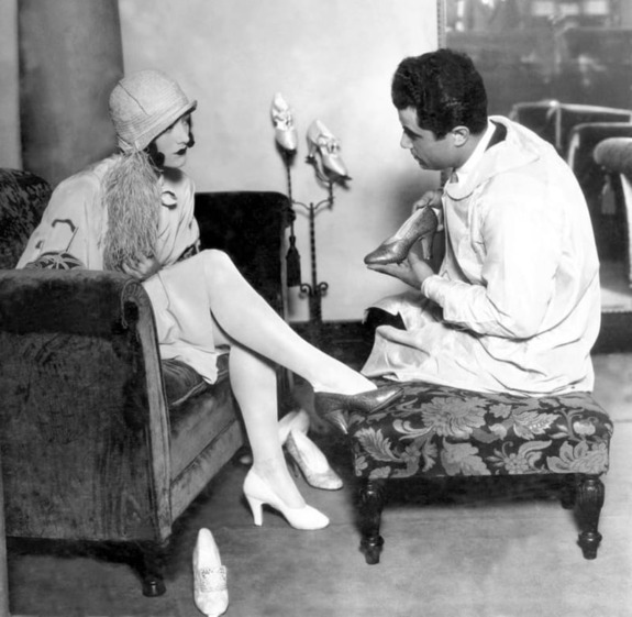 Woman trying on shoes with help from a shoemaker in a vintage setting.