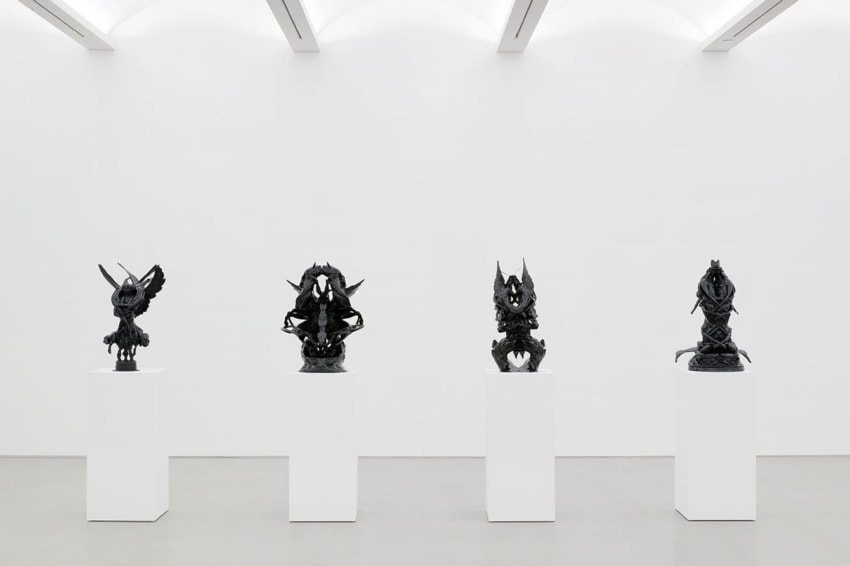 Four intricate black sculptures displayed on white pedestals in a modern gallery with a minimalist design.