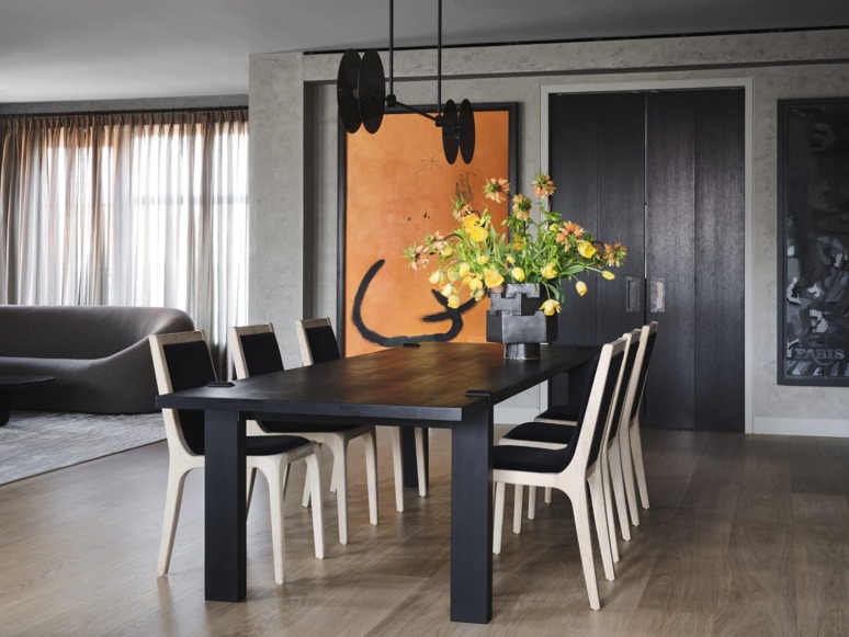 Modern dining room with a black table, wooden chairs, vibrant yellow flowers, large orange artwork, and soft lighting.