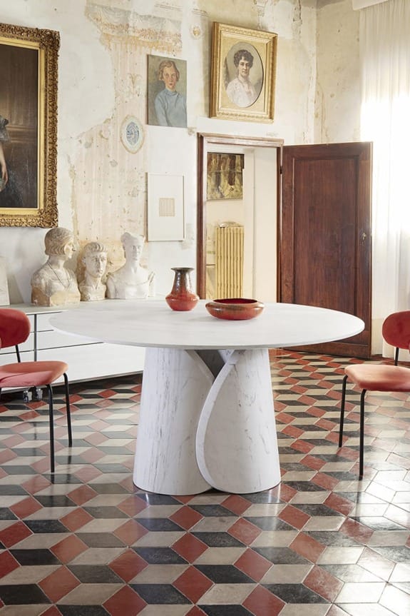 Round marble table with decorative items in an elegant room featuring statues, vintage paintings, and a checkered floor.