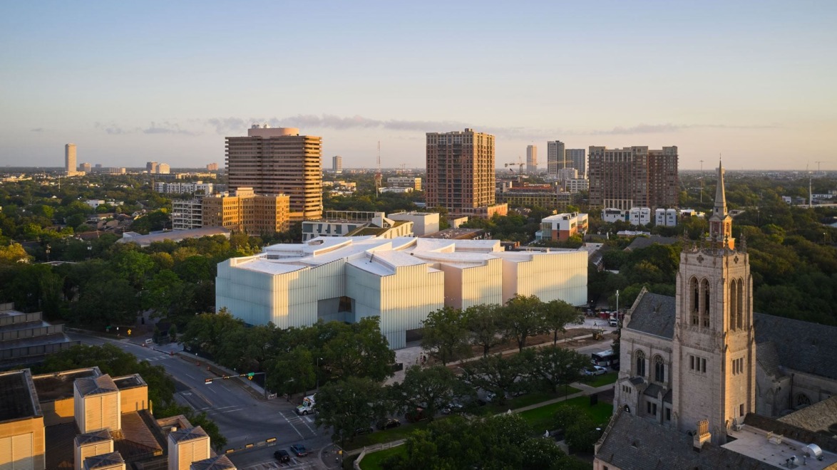 Aerial view of modern glass building surrounded by trees and cityscape at sunset.
