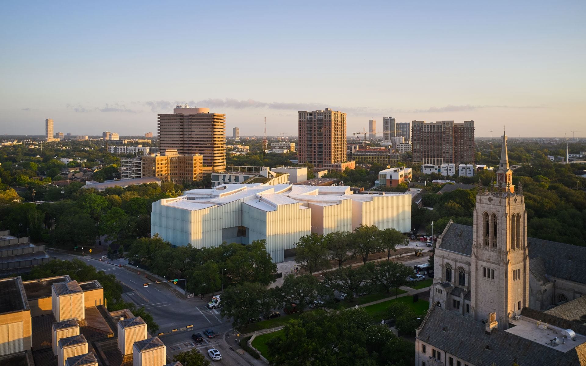 Aerial view of modern glass building surrounded by trees and cityscape at sunset.