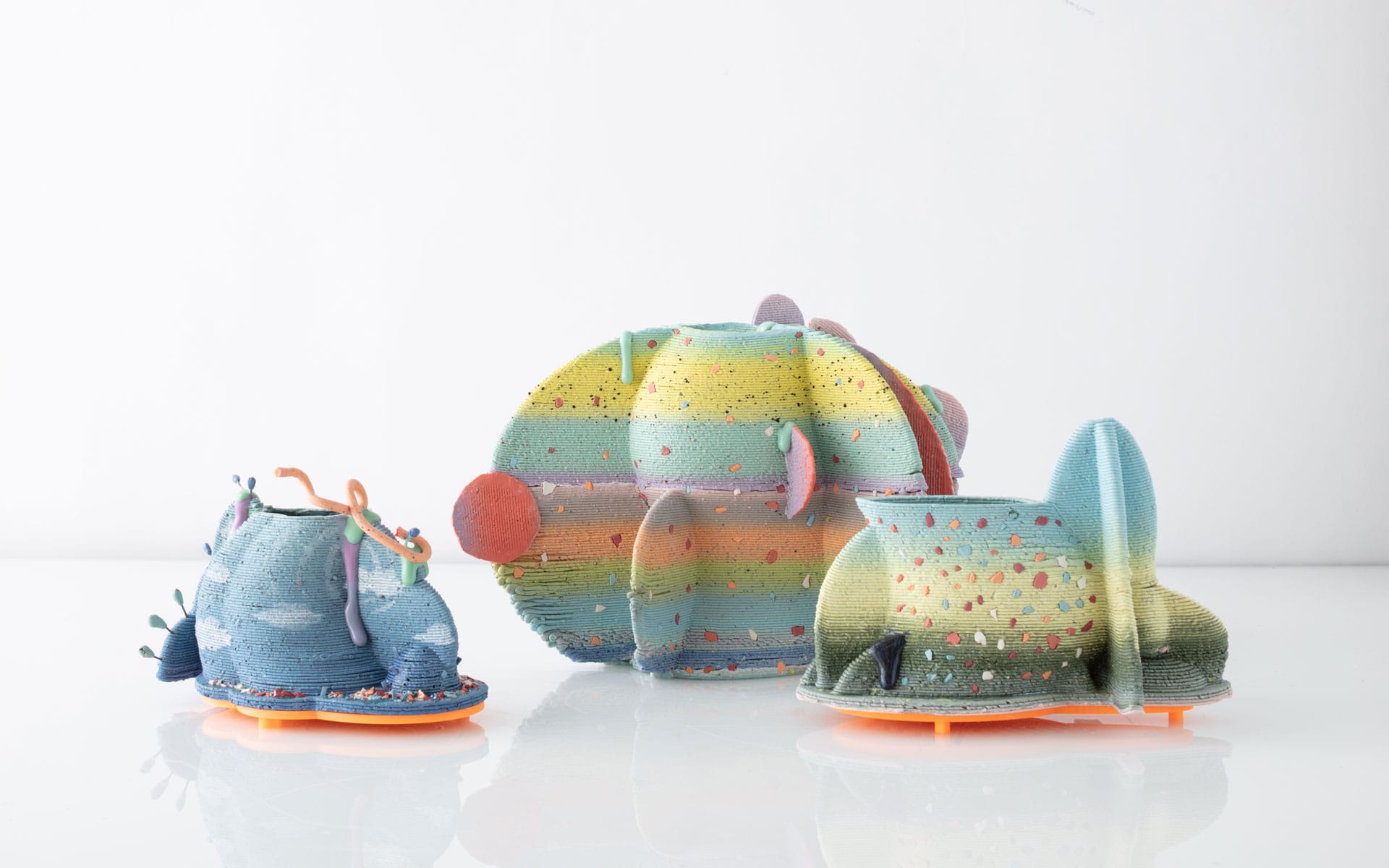 Colorful, abstract 3D printed sculptures in various shapes and sizes on a reflective white surface.