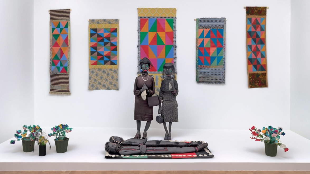 Two sculptures facing each other in a gallery, surrounded by colorful geometric textiles and small flower arrangements.