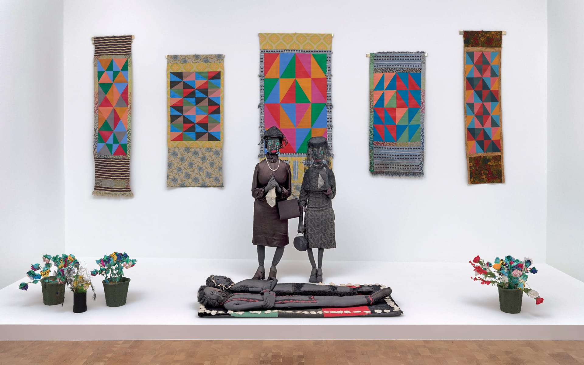 Two sculptures facing each other in a gallery, surrounded by colorful geometric textiles and small flower arrangements.