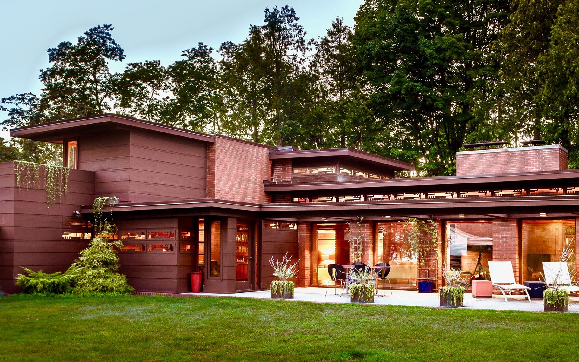 Modern house with large windows and flat roofs, surrounded by trees, featuring a spacious patio with outdoor seating.