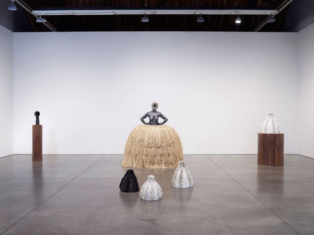 Art installation featuring sculptures on pedestals and floor, central figure with straw base in a gallery with white walls.