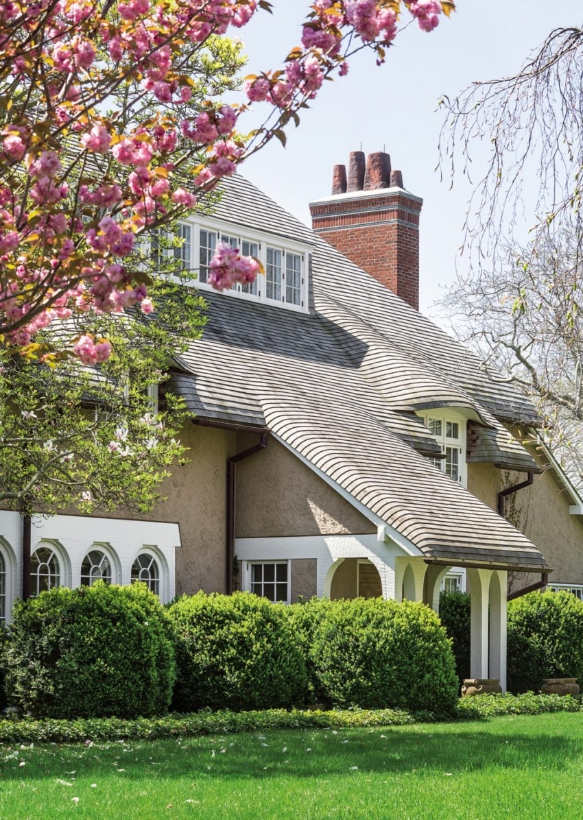 Charming cottage with a sloped roof surrounded by lush greenery and blossoming trees under a clear blue sky.