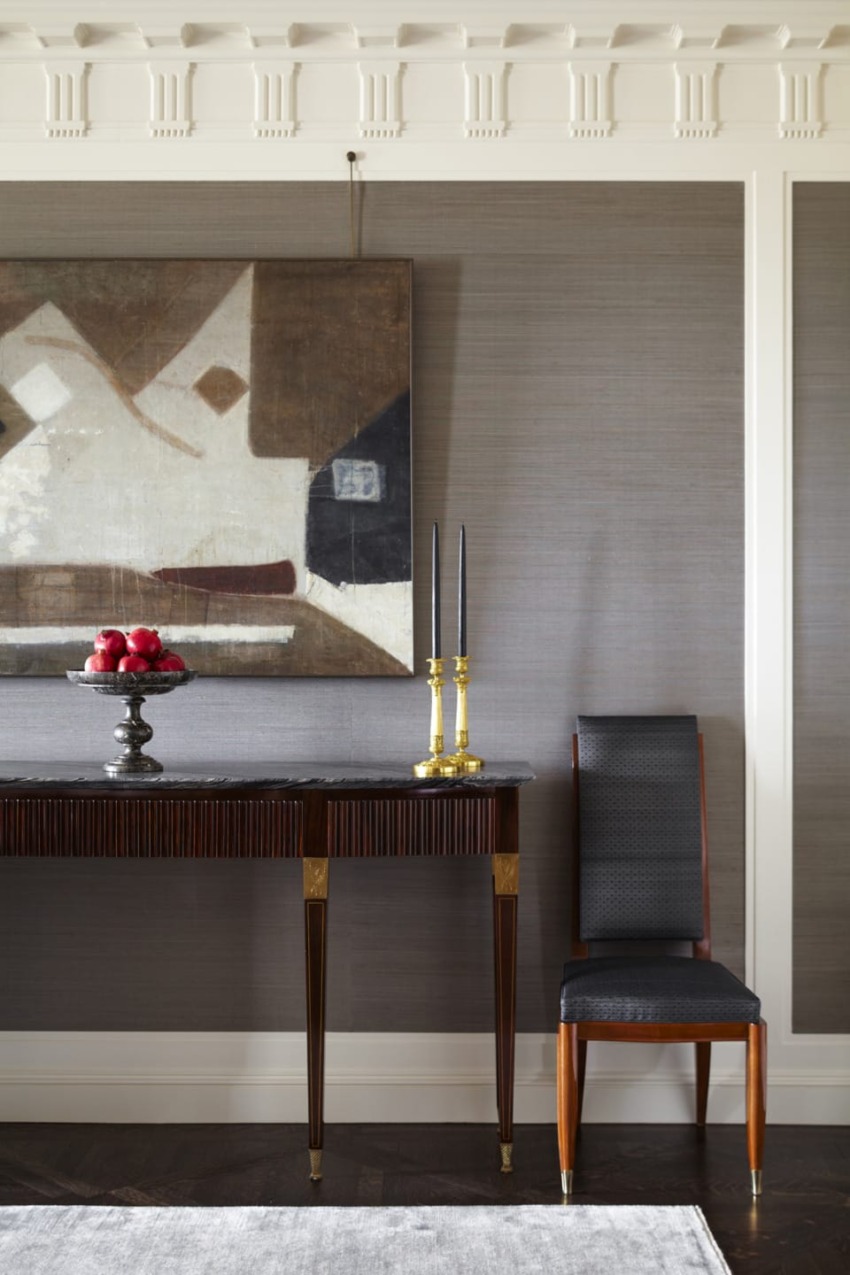 Elegant room with abstract painting, black candlesticks, fruit bowl on a table, and a black-cushioned chair against gray walls.