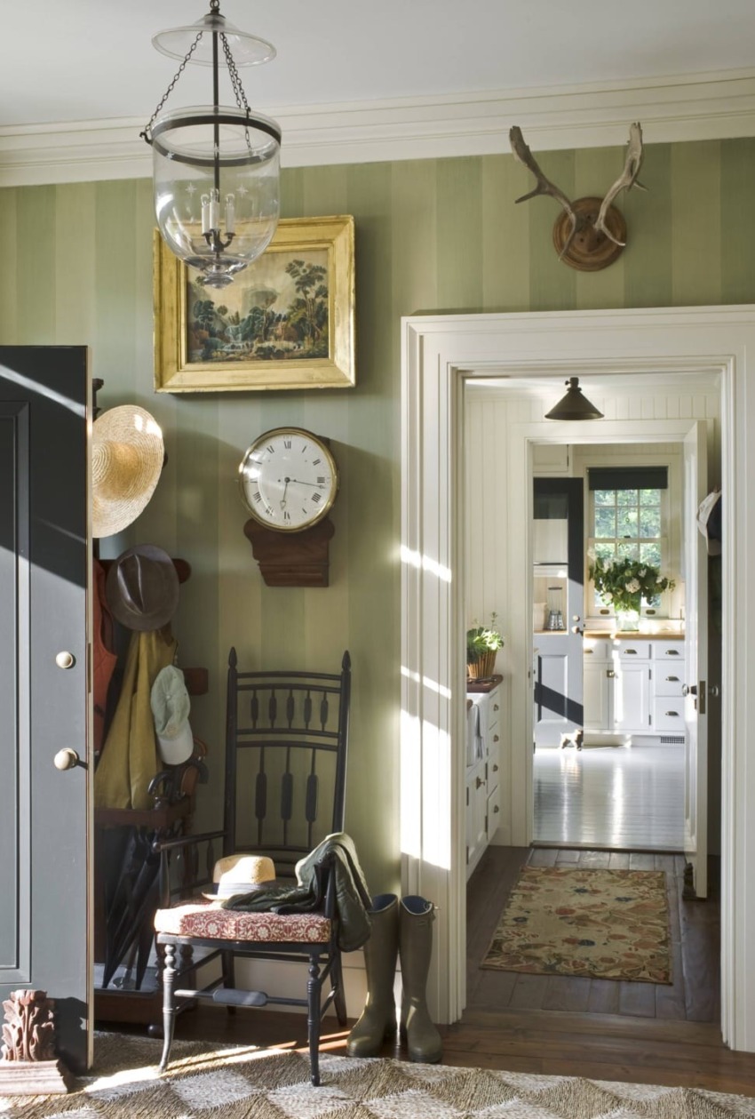 Entryway with vintage decor, hats on hooks, a clock, a chair, and a view into a bright kitchen with plants.