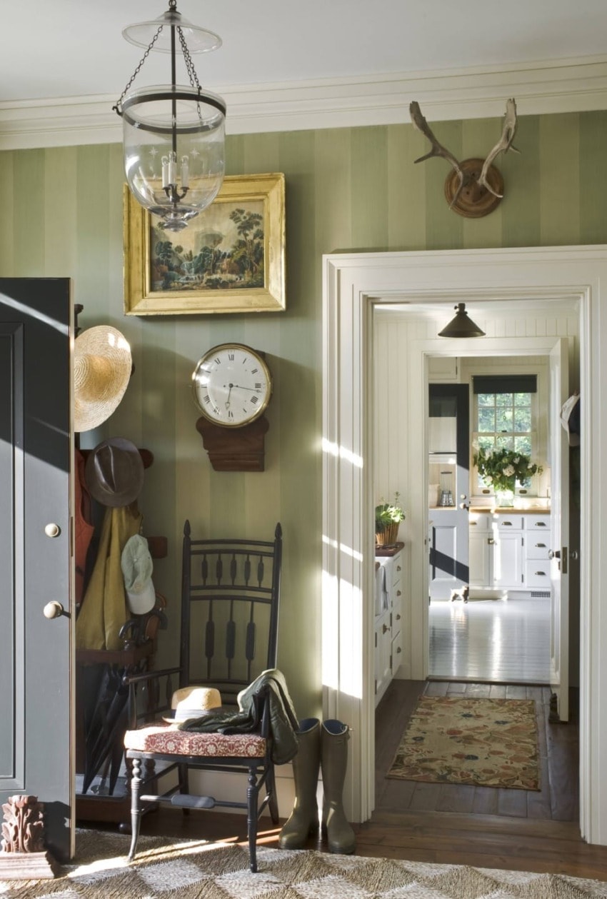 Entryway with vintage decor, hats on hooks, a clock, a chair, and a view into a bright kitchen with plants.