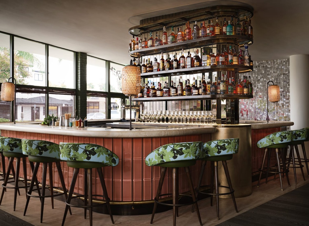 Modern bar with green floral stools, curved marble counter, and shelves filled with bottles against large windows.