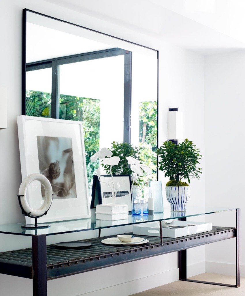 Modern living room with a glass table, decorative items, large framed mirror, and potted plants near a window.